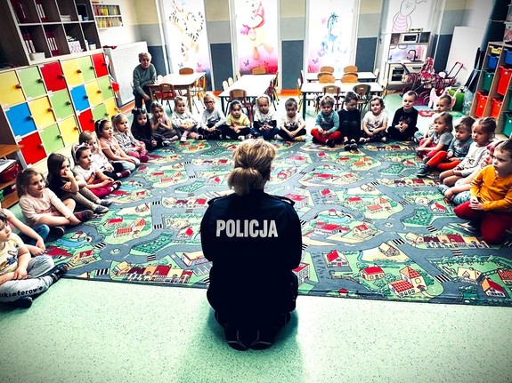 policjantka siedząca tyłem do obiektywu przemawiająca do dzieci siedzących na dywaniku