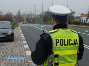 Policjant z ruchu drogowego w kazmizelce odblaskowej stoi przy jezdni podczas akcji związanej z bezpieczeństwem na drodze